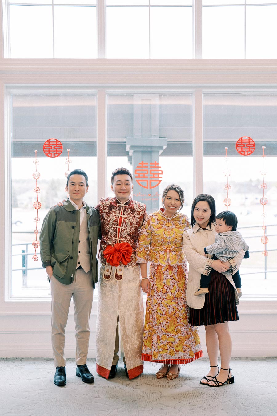 A group of people celebrating a traditional Chinese event, with two individuals in ornate traditional attire featuring intricate red and gold designs. They are joined by three others in modern clothing, standing in front of a window adorned with Chinese festive decorations.