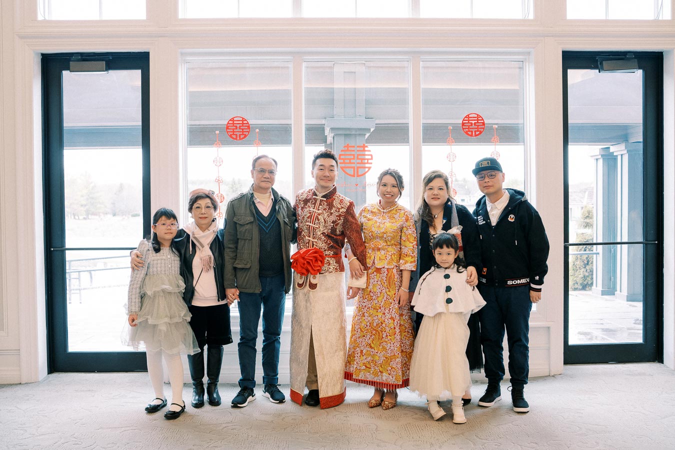 A family standing together in front of glass doors, featuring two people in traditional Chinese wedding attire.