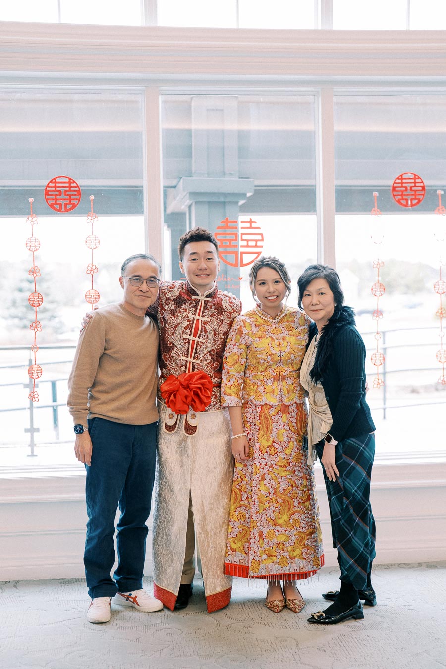 A family posing together at a traditional Chinese wedding, with decorative red symbols and patterns on the window in the background. The couple is wearing ornate, colorful wedding attire, featuring intricate embroidery and designs, while standing indoors in a brightly lit room.