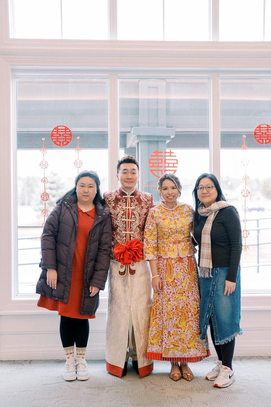 A group of four people standing indoors in front of a window, with two people wearing traditional Chinese wedding attire and two others in casual clothing. Red Chinese decorations adorn the window.