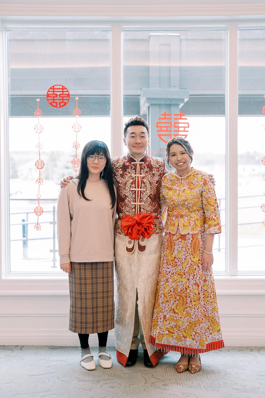Three people posing in traditional Chinese wedding attire, with two wearing elaborate embroidered outfits, standing in front of a window decorated with red Chinese symbols, suggesting a cultural celebration or ceremony.