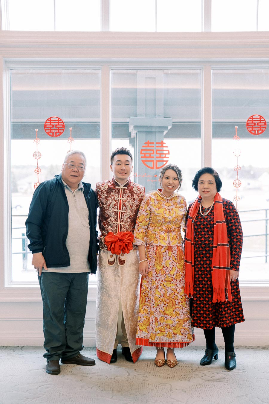 A family photo during a traditional cultural celebration, featuring a couple in ornate ceremonial attire with red accents, standing alongside two relatives inside a decorated room.