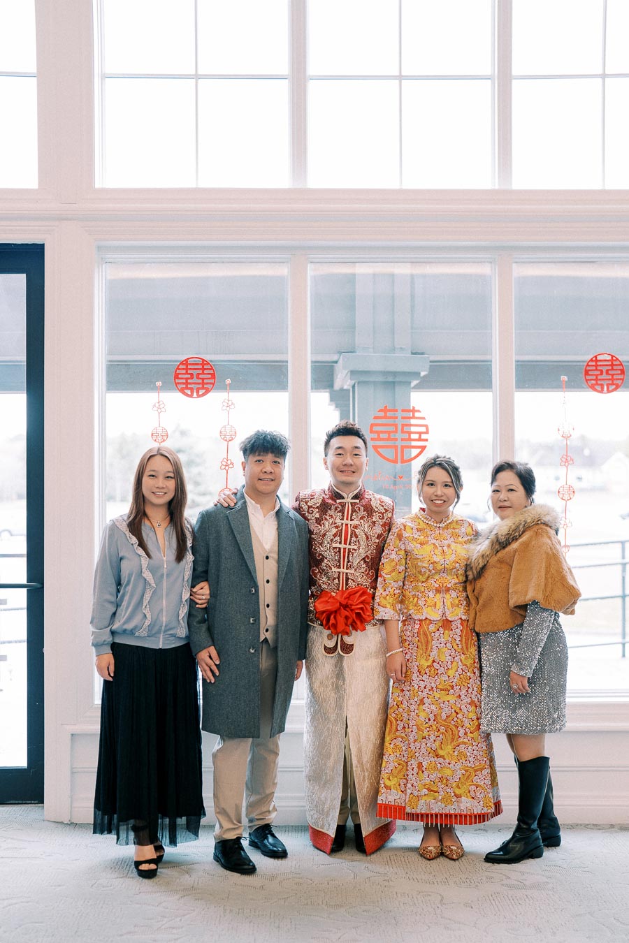 Family posing for a portrait at a cultural event, featuring two individuals in traditional Chinese attire and three others in modern formal clothing, standing in front of a window with decorative red symbols.