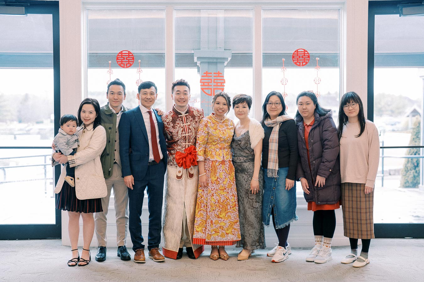 A group of people smiling for a family photo at a traditional wedding ceremony, featuring the couple in elegant traditional attire, surrounded by family members against a bright window backdrop with festive decorations.
