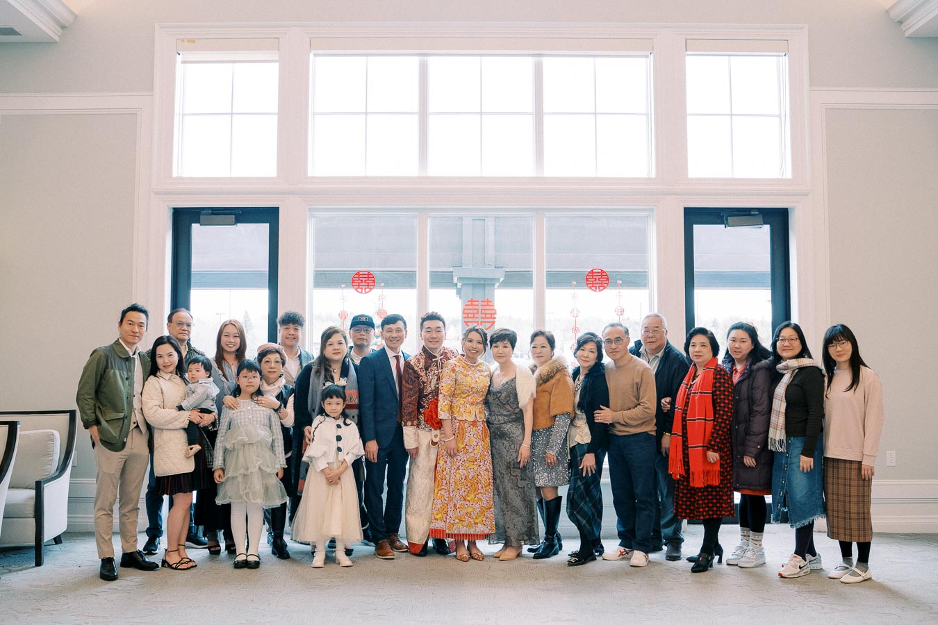 A large family group poses indoors in front of bright windows, with a couple dressed in traditional Chinese attire at the center, symbolizing a cultural celebration or family gathering.