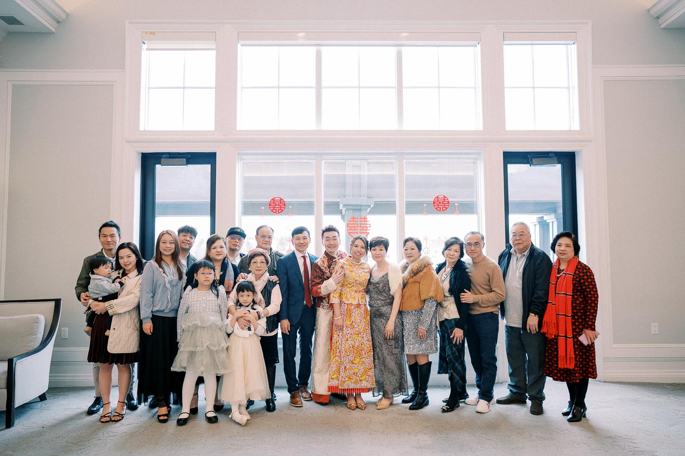 A large family group posing together indoors at a celebration, with a couple in traditional attire at the center, surrounded by relatives of various ages including children, in front of a window decorated with red festive symbols.