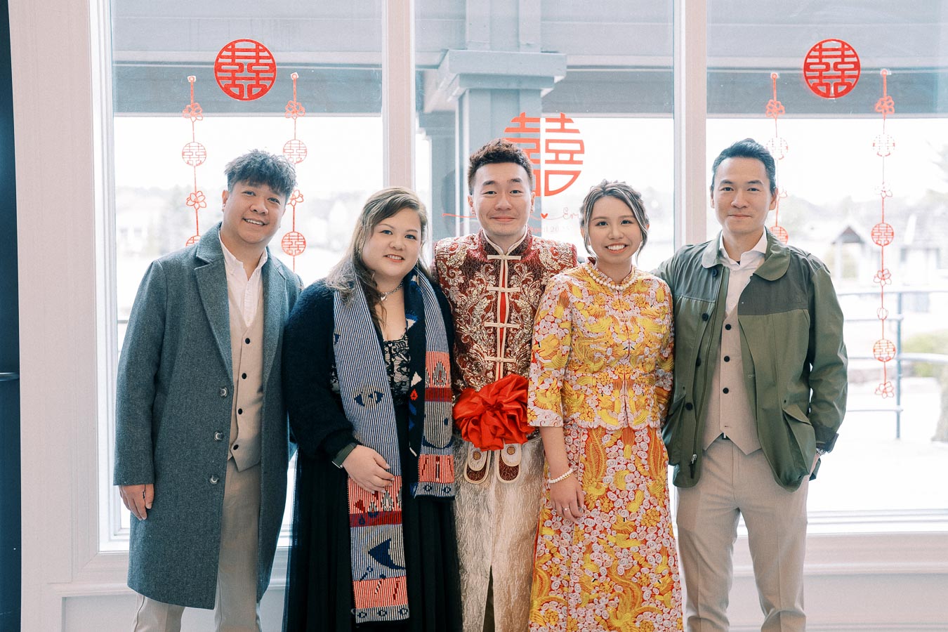 A group of people dressed in formal attire standing in front of a window with red Chinese decorative symbols. The individuals in the center are wearing traditional ceremonial clothing, suggesting a cultural celebration or event.