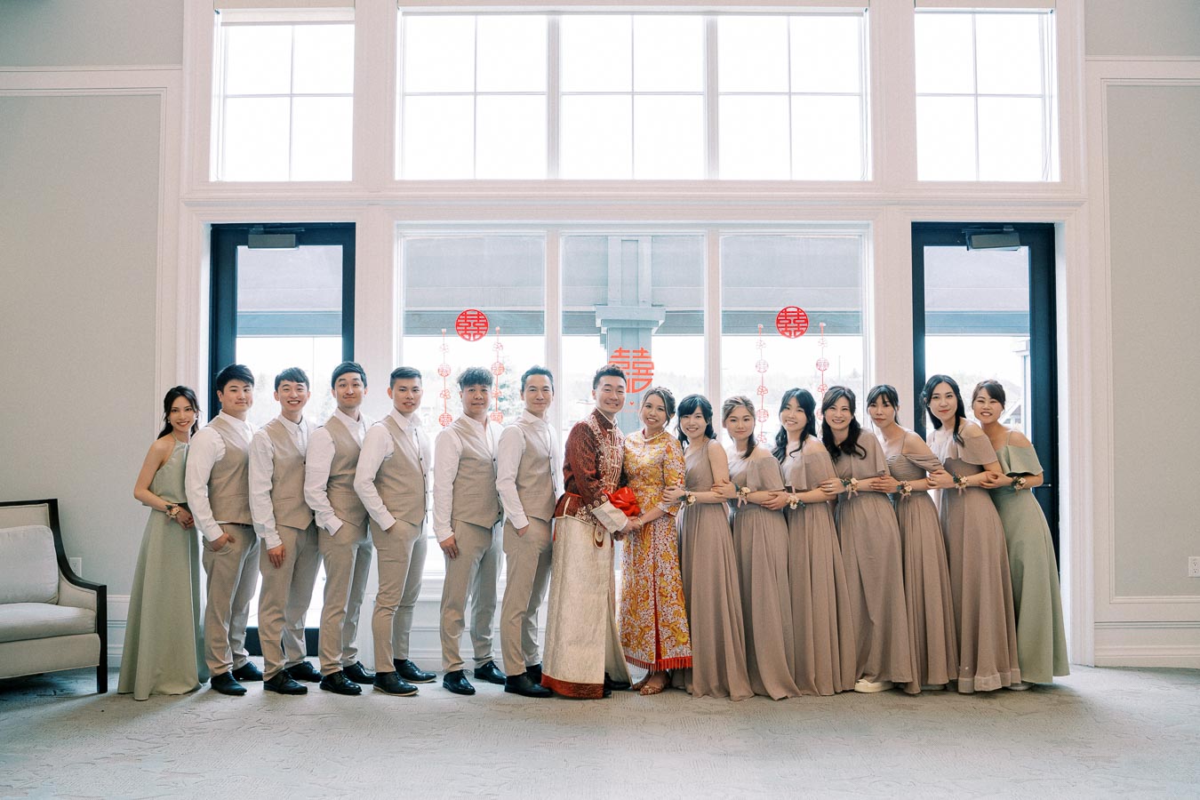 A joyful wedding party featuring traditional attire, with the bride and groom surrounded by bridesmaids in taupe dresses and groomsmen in beige vests, posing in front of large windows adorned with red festive decorations.