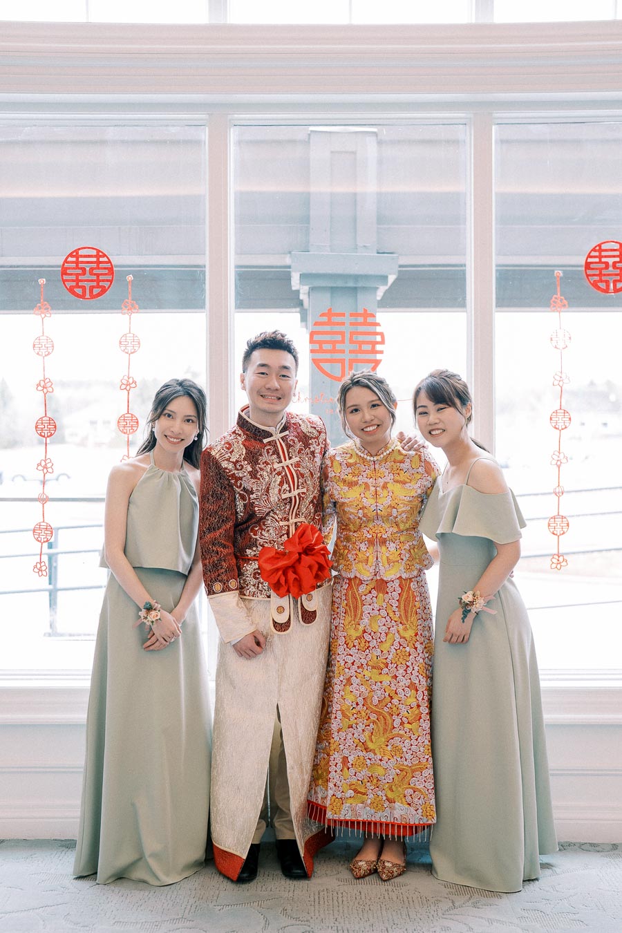 A wedding couple in traditional Chinese attire stands with two bridesmaids in pastel dresses, posed in front of a window decorated with red Chinese symbols.