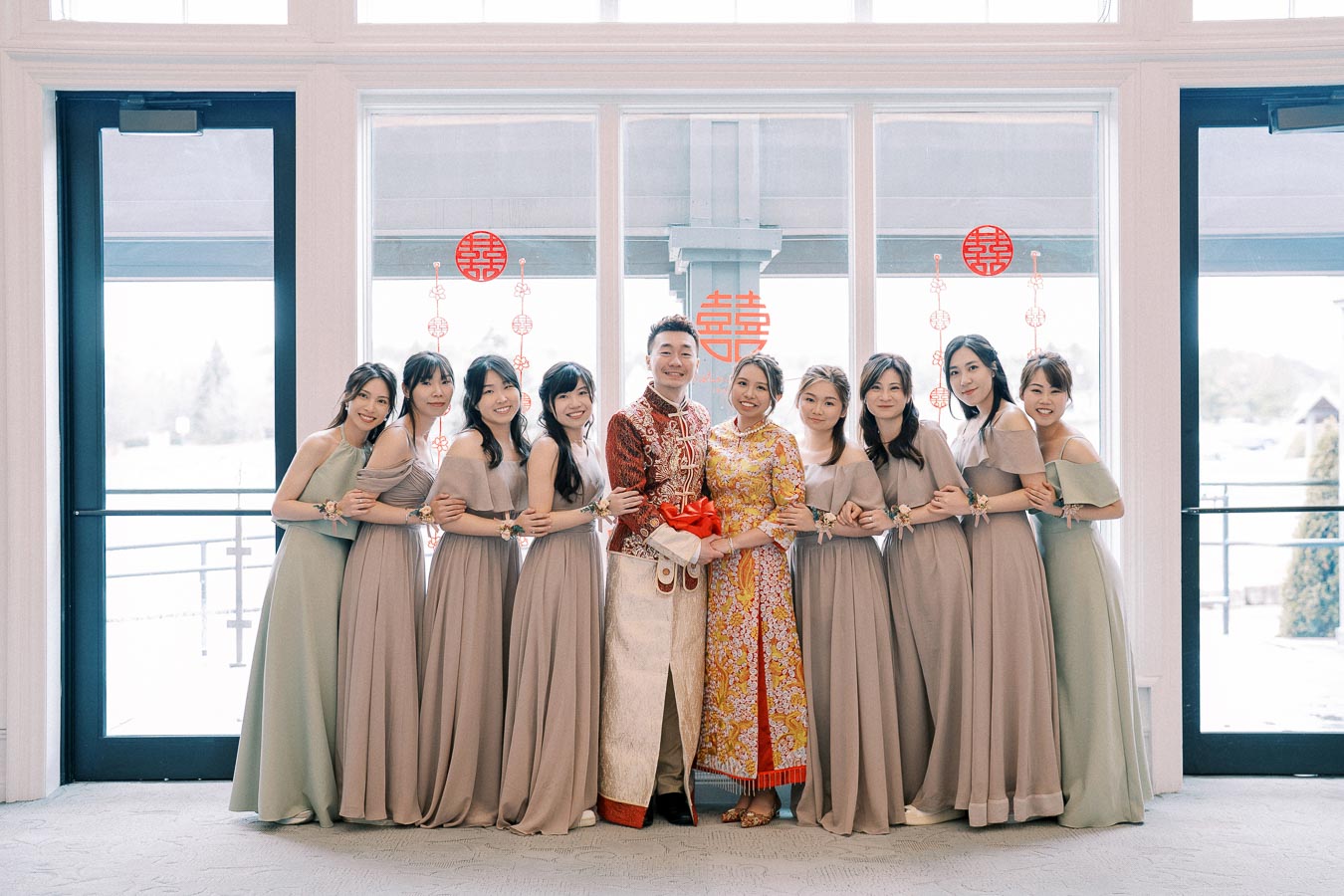 Traditional Chinese wedding couple surrounded by bridesmaids in elegant gray dresses, standing in front of a window with red Chinese double happiness symbols.