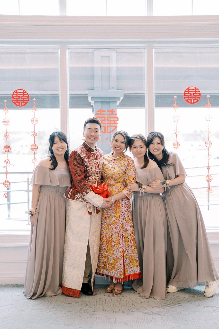 A joyful couple in traditional Asian wedding attire surrounded by bridal party members in gray dresses, standing in front of a window decorated with red Chinese characters for double happiness.