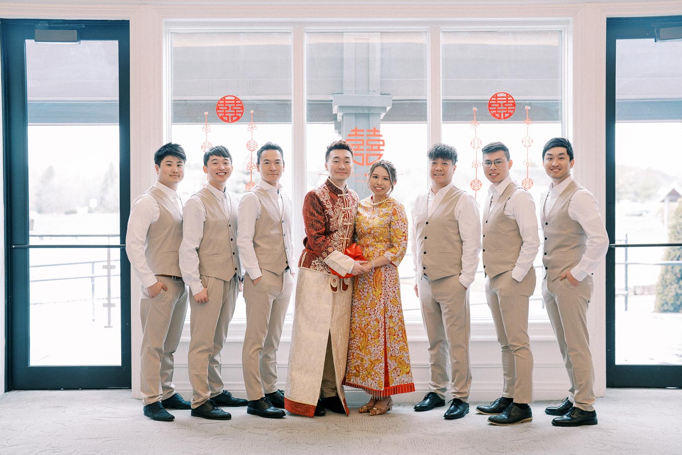 Asian couple in traditional wedding attire surrounded by their wedding party in beige suits, standing near a window with Chinese double happiness decorations.
