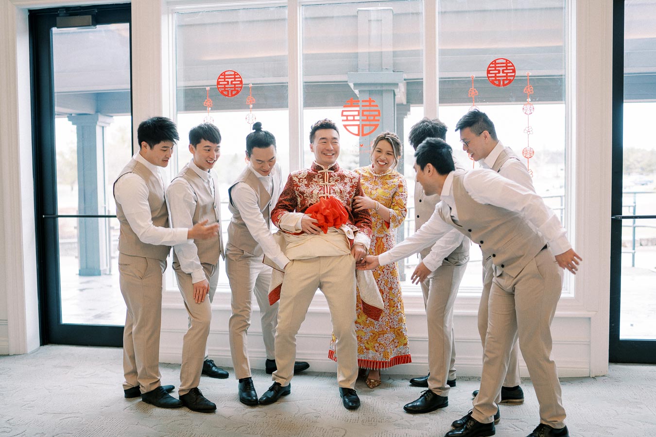 A joyful group of people celebrating a traditional Chinese wedding, with the groom dressed in ornate red attire, surrounded by friends in light-colored suits and the bride in a colorful dress, symbolizing happiness and unity.