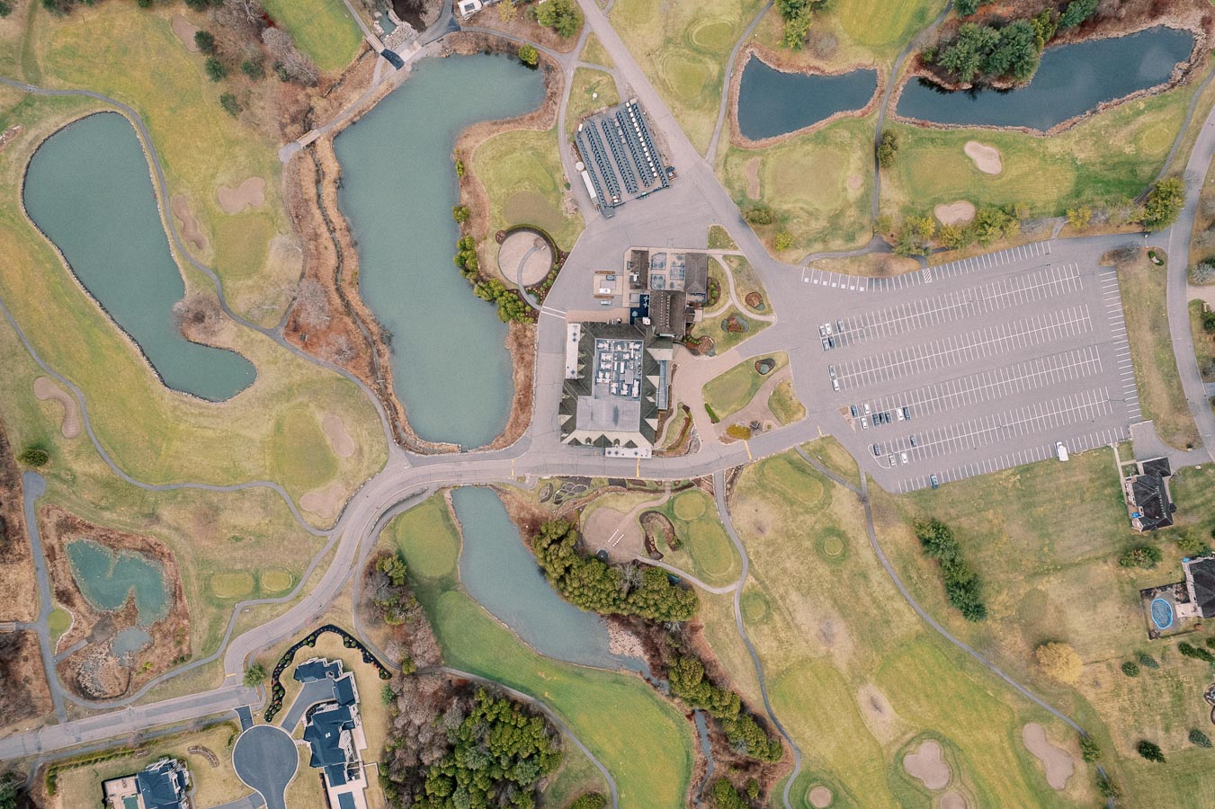 Aerial view of a lush golf course with multiple ponds, a central clubhouse, and a large parking area. The landscape features winding paths and well-maintained greens surrounded by trees and residential areas. Perfect for a relaxing day of golfing.