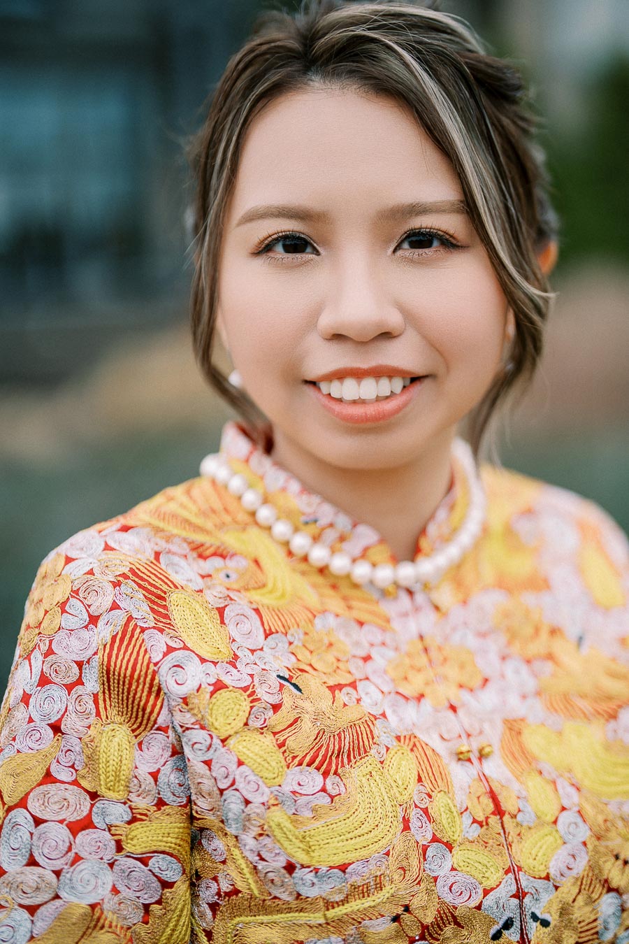 Smiling person wearing an ornate, colorful traditional outfit with intricate embroidery, and a pearl necklace, outdoors.