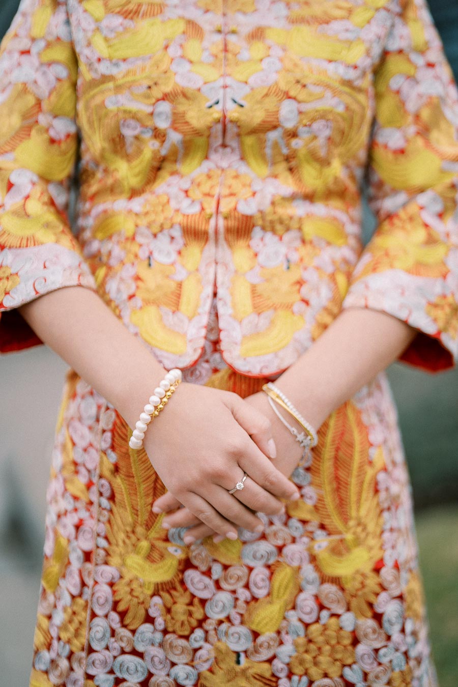 A close-up of a person wearing a colorful, intricately patterned traditional dress with floral designs. The hands are adorned with elegant pearl bracelets and a delicate ring, highlighting cultural fashion and accessories.