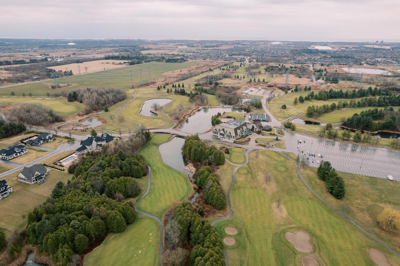 Aerial view of a lush golf course with well-maintained greens, sand traps, and winding pathways surrounded by residential houses and a large clubhouse. The landscape is complemented by small lakes and patches of dense trees under an overcast sky.