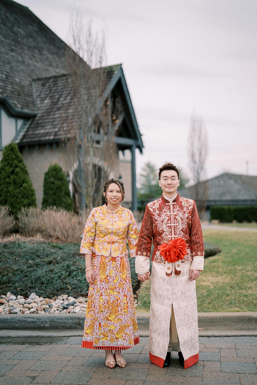 A couple in traditional Chinese attire posing outdoors in front of a building, with the woman wearing a colorful floral dress and the man in an ornate red and white outfit.