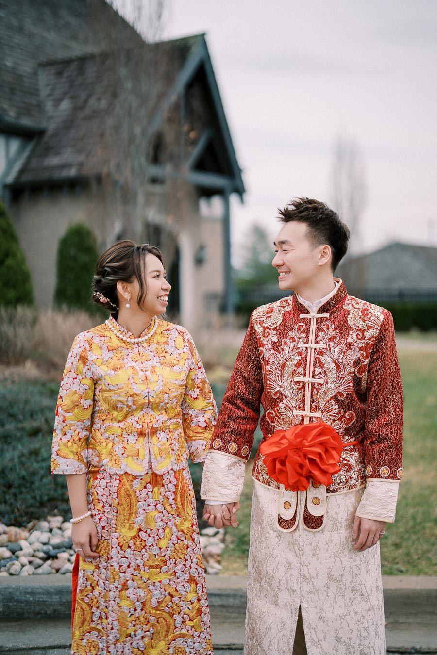 A couple in traditional Asian wedding attire holding hands and smiling at each other outside a building with a garden background.