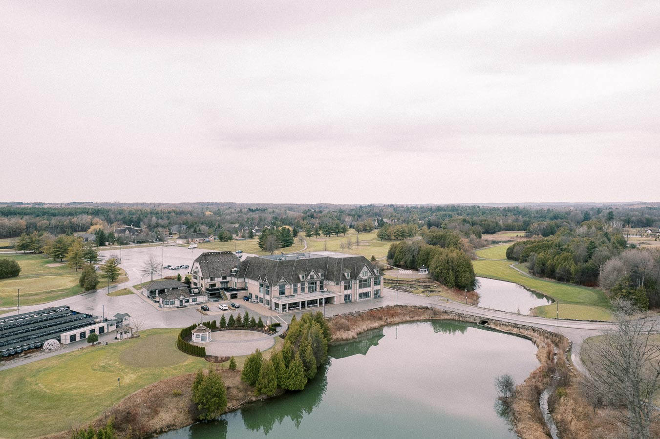 Aerial view of a large countryside property featuring a grand building with a sloped roof, surrounded by lush greenery and a tranquil pond on a cloudy day.