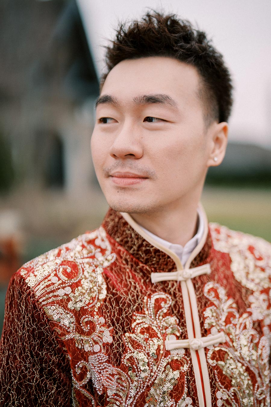 Portrait of a man wearing a traditional red and gold embroidered outfit, looking to the side outdoors.
