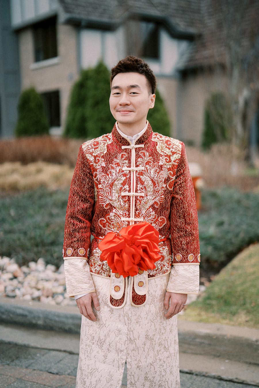 Portrait of a person wearing a traditional red and gold embroidered outfit with a large red decorative bow, standing outdoors in front of a residential building.