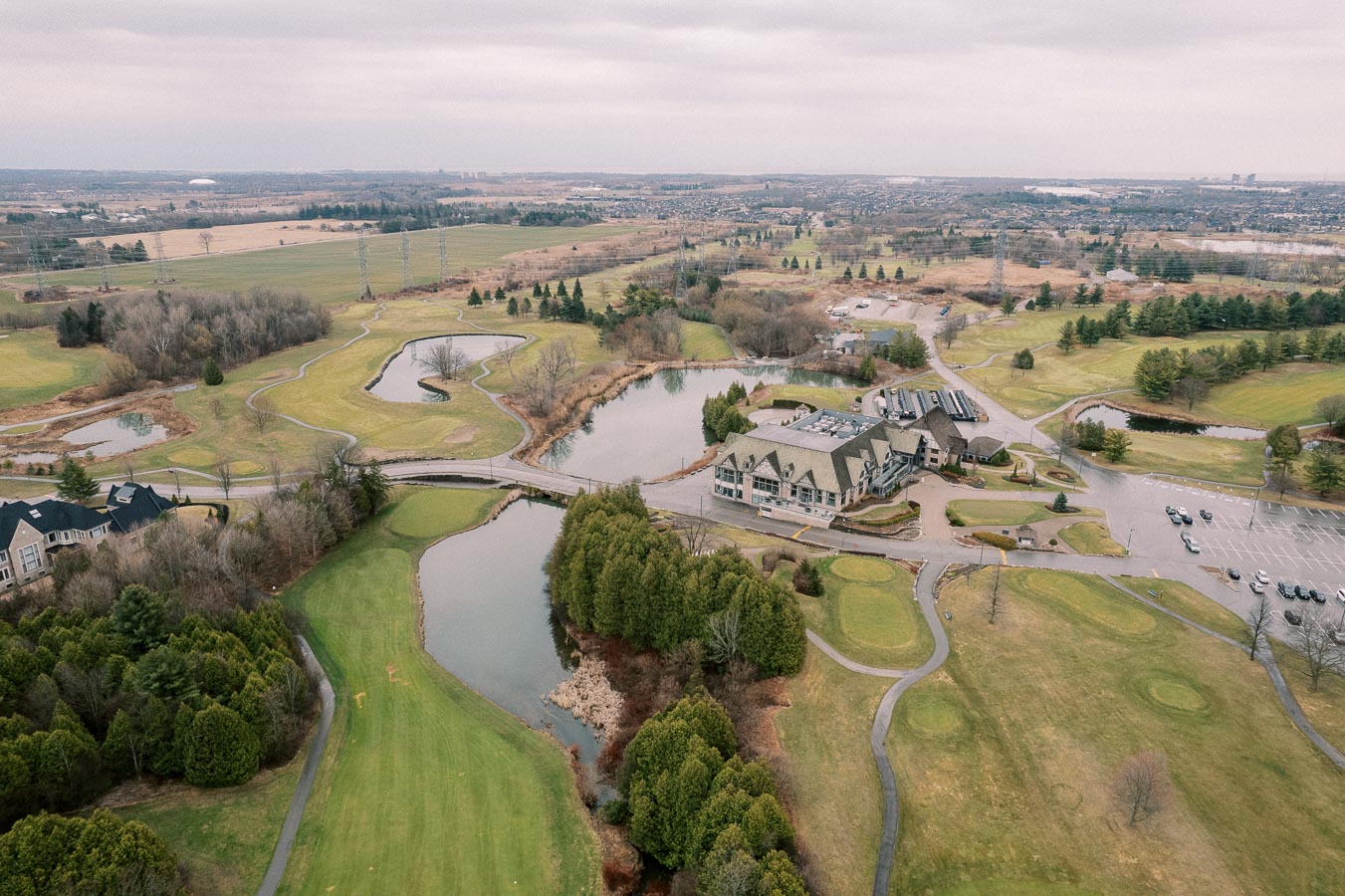 Aerial view of a sprawling golf course featuring lush green fairways, picturesque water hazards, and a clubhouse with surrounding parking area, set in a serene landscape with distant hills and city skyline in the background.