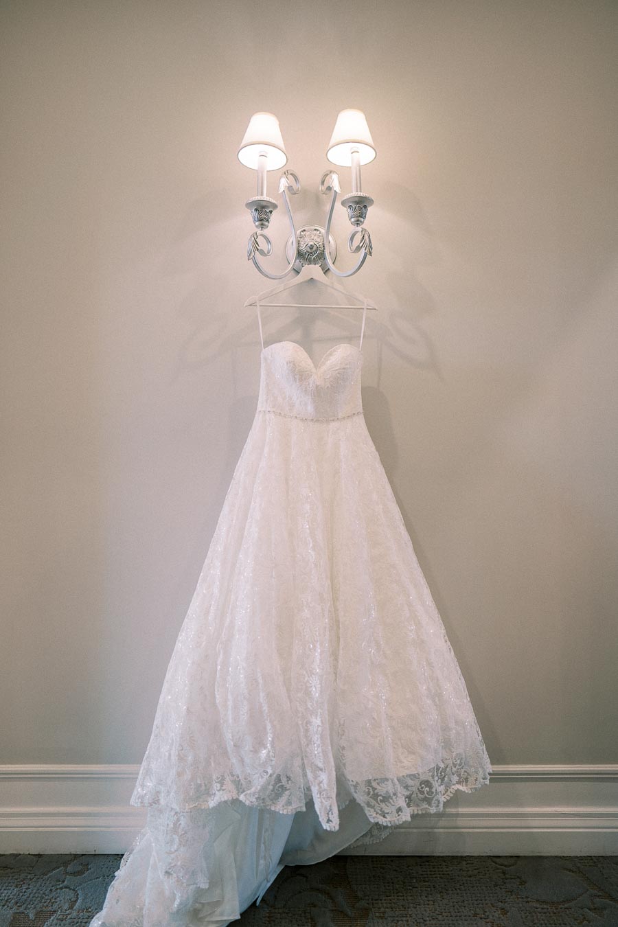 Elegant white lace wedding dress hanging on wall beneath a stylish light fixture.