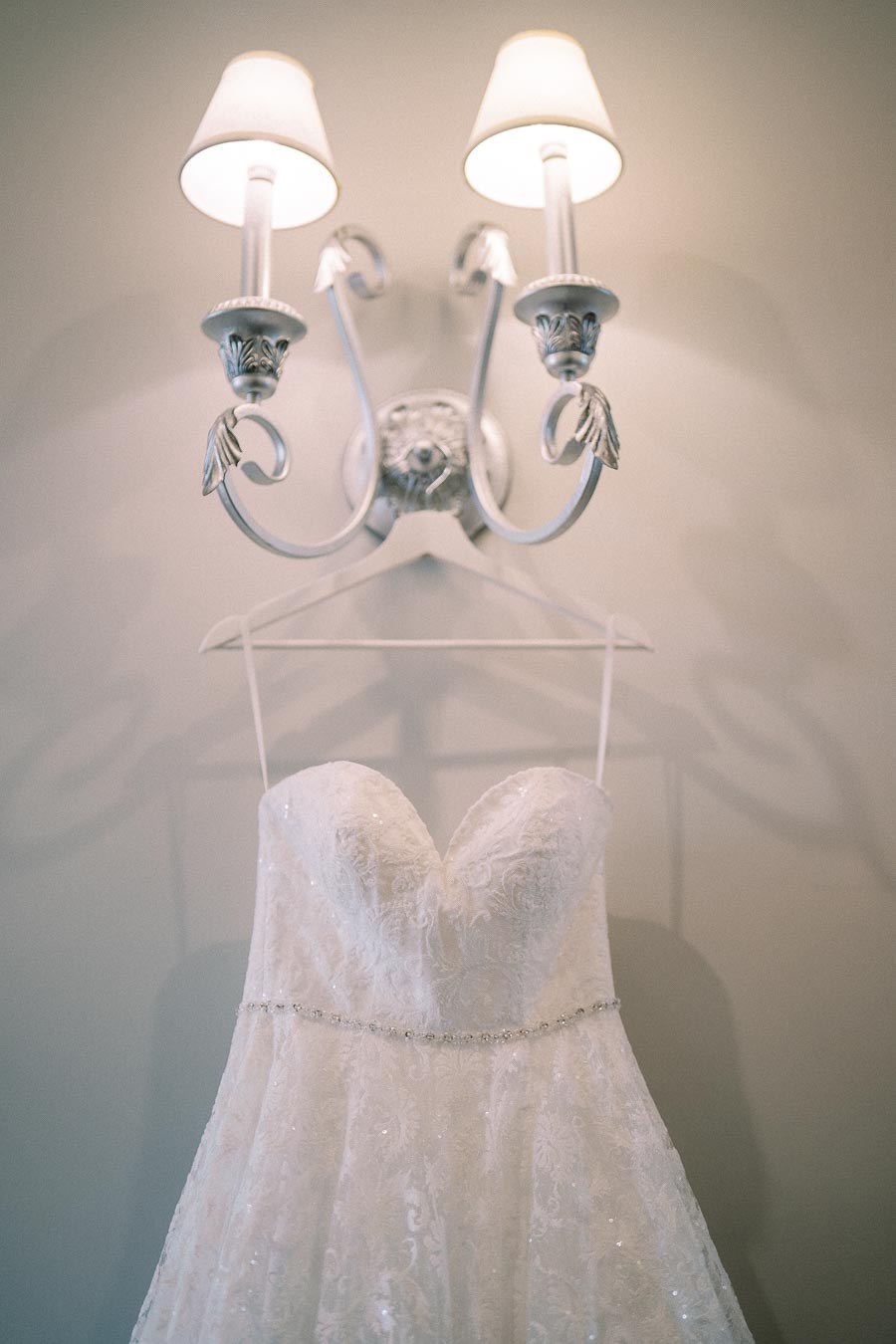 Elegant strapless white lace wedding dress hanging under vintage style wall sconce with soft lighting.
