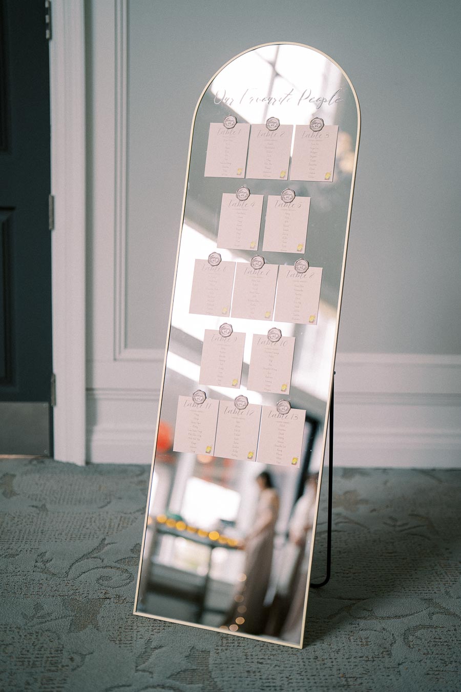 Wedding seating chart displayed on a tall mirror, featuring elegantly arranged table assignments with calligraphy, set in a sophisticated venue.