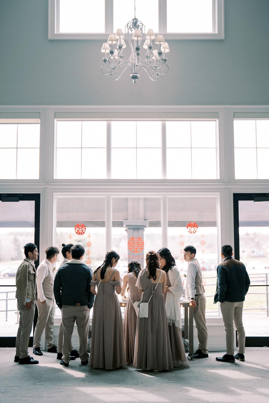 A group of people in formal attire gather around a large table in a well-lit room with tall windows and a chandelier, decorated with traditional red Chinese symbols.