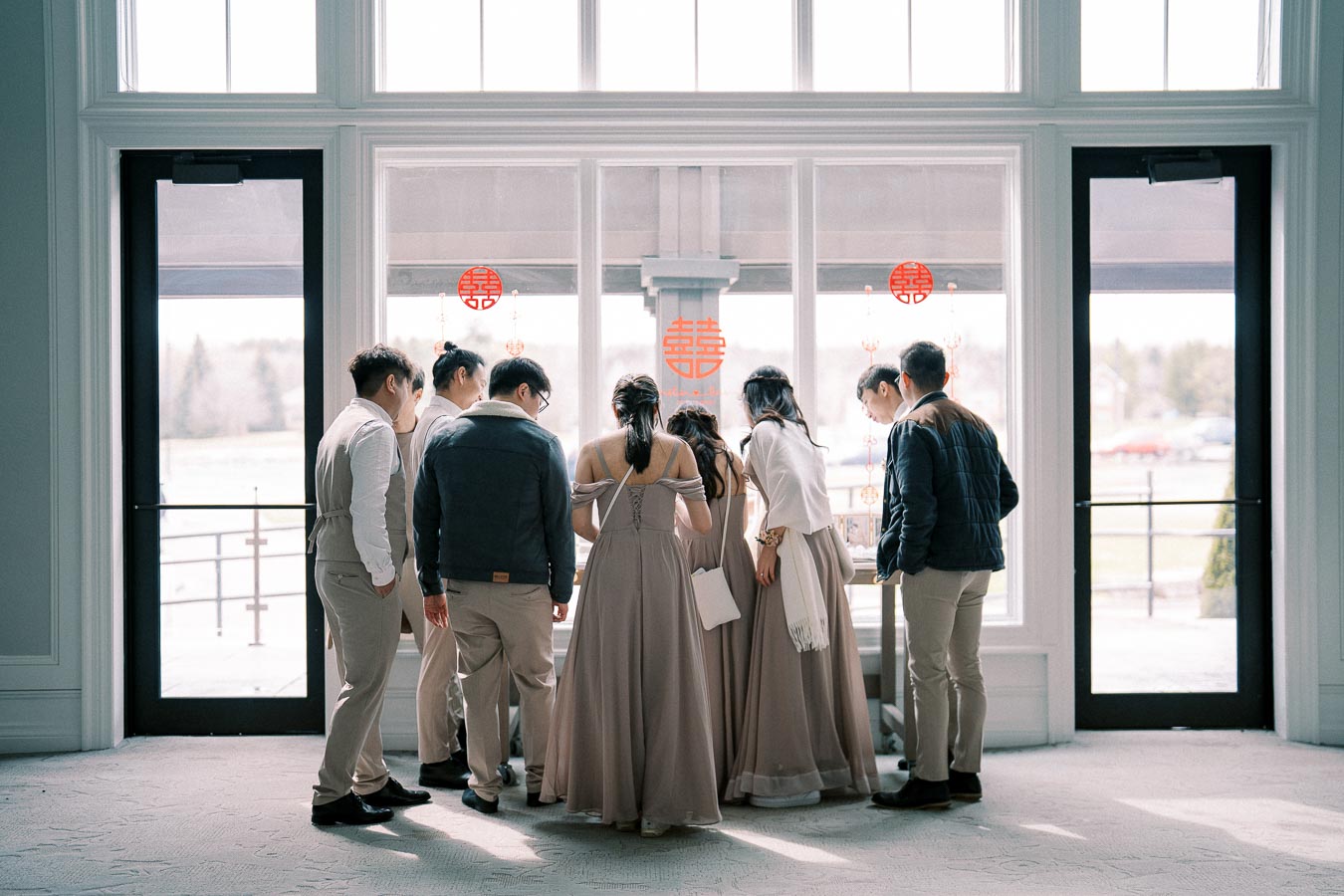 A group of elegantly dressed people gather around a window adorned with red double happiness symbols, often associated with weddings, suggesting a celebratory or formal event setting. The soft lighting and serene atmosphere enhance the occasion's sophistication.