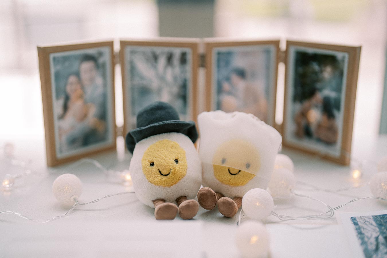 Two smiling egg-themed plush toys, one wearing a black hat, sit in front of a folding picture frame with blurred photographs in the background. String lights are artfully placed around them, creating a cozy atmosphere.