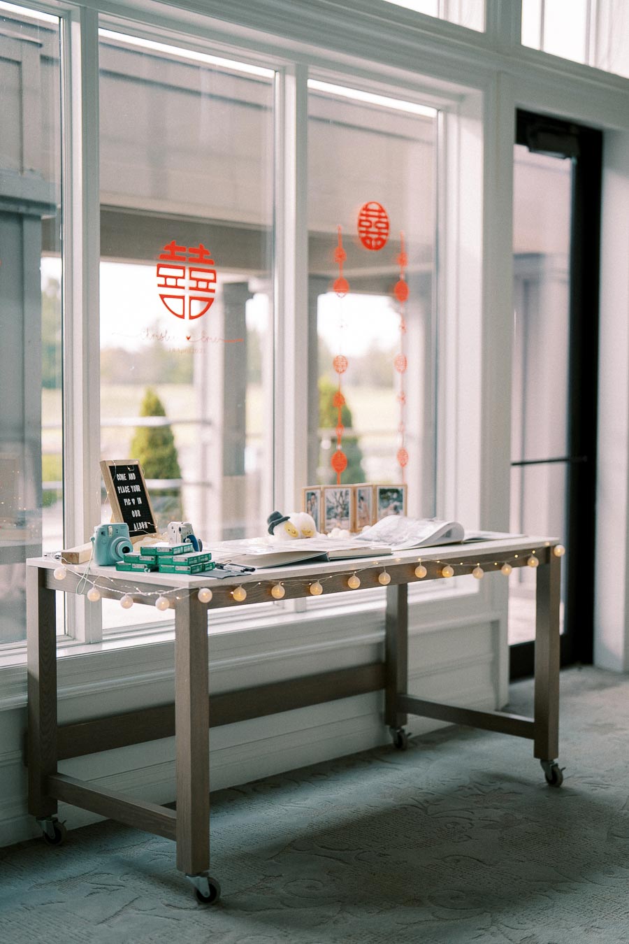 A decorative table setup inside a bright room with large windows featuring red Chinese wedding symbols, adorned with string lights, instant cameras, and guestbooks for a wedding reception.