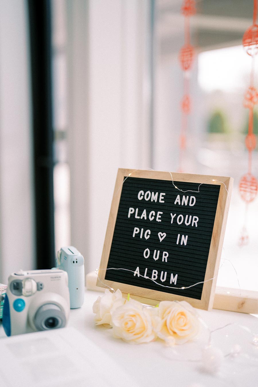 A decorative display with a letter board inviting guests to Come and place your pic in our album, surrounded by white roses, a polaroid camera, and string lights, creating an inviting and festive atmosphere.