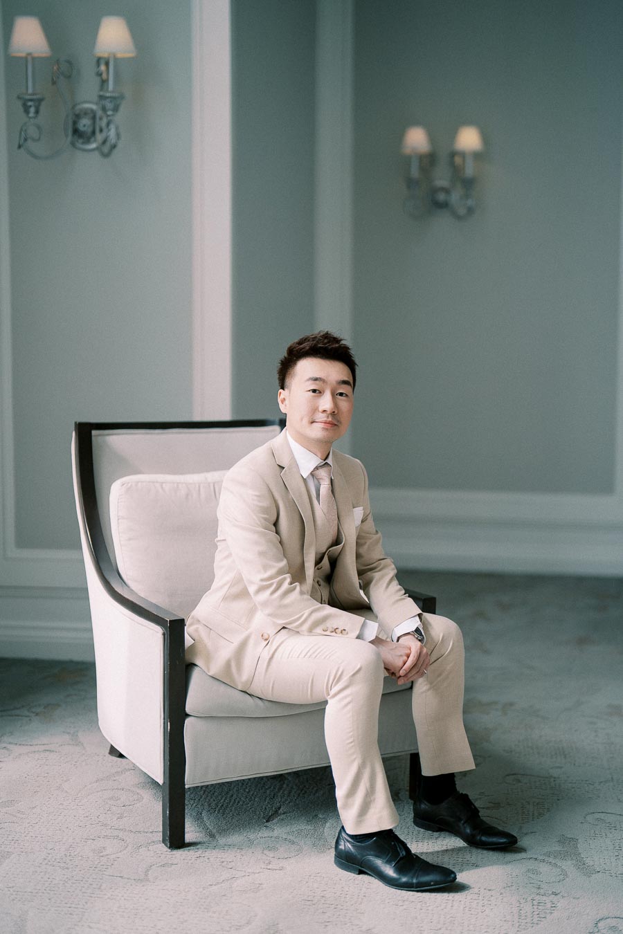 A man in a beige suit sits confidently on a chair in a well-lit, elegant room with light-colored walls and decorative sconces.