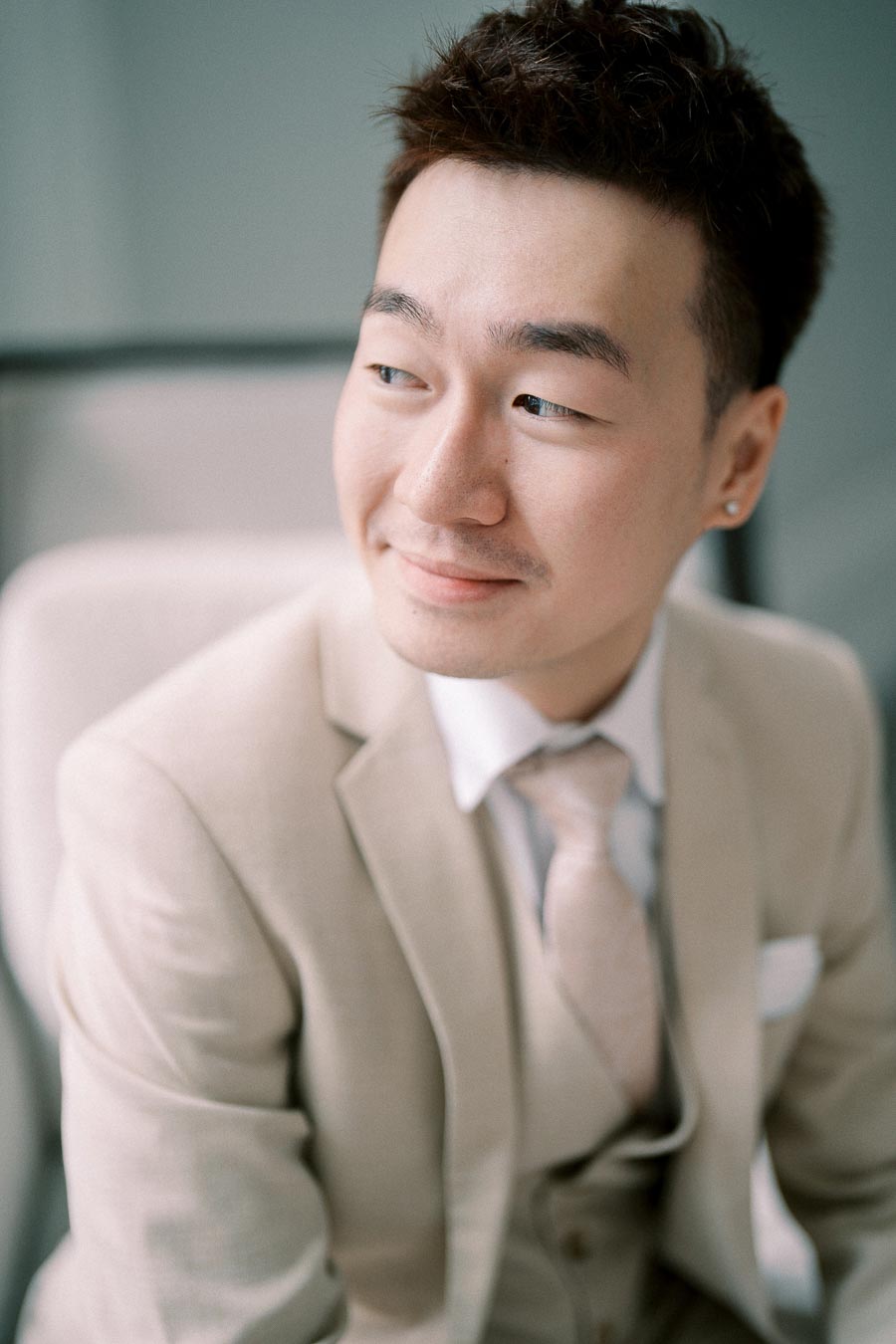 A man in a beige suit and tie looking to the side with a relaxed expression, seated on a chair in a softly lit modern environment.
