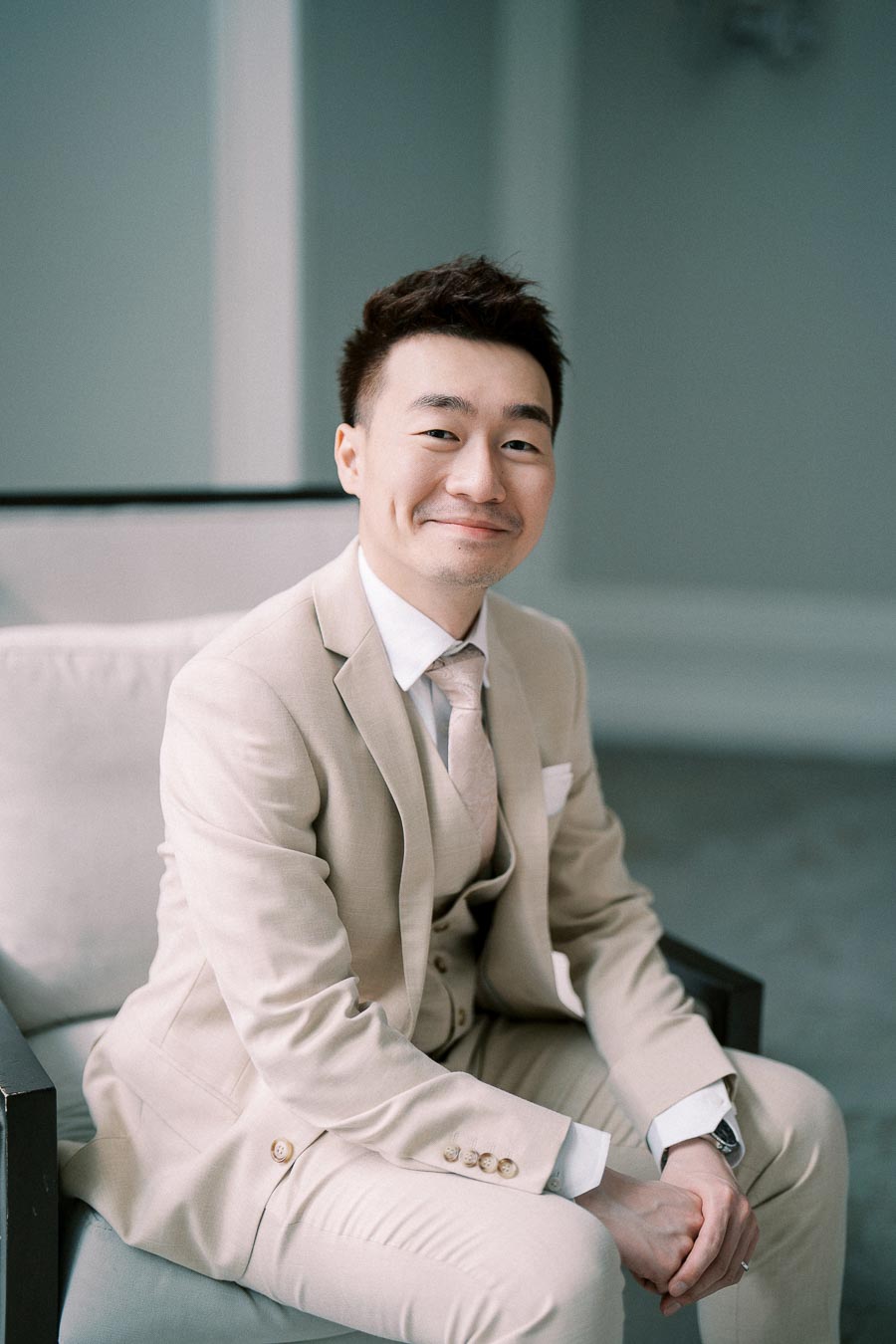 Man in a light beige suit seated on a chair, smiling at the camera in a softly lit room.