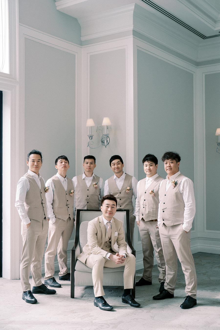 A stylish group of seven groomsmen in beige suits with coordinated floral boutonnieres, posing elegantly in a bright, sophisticated room with classic decor.