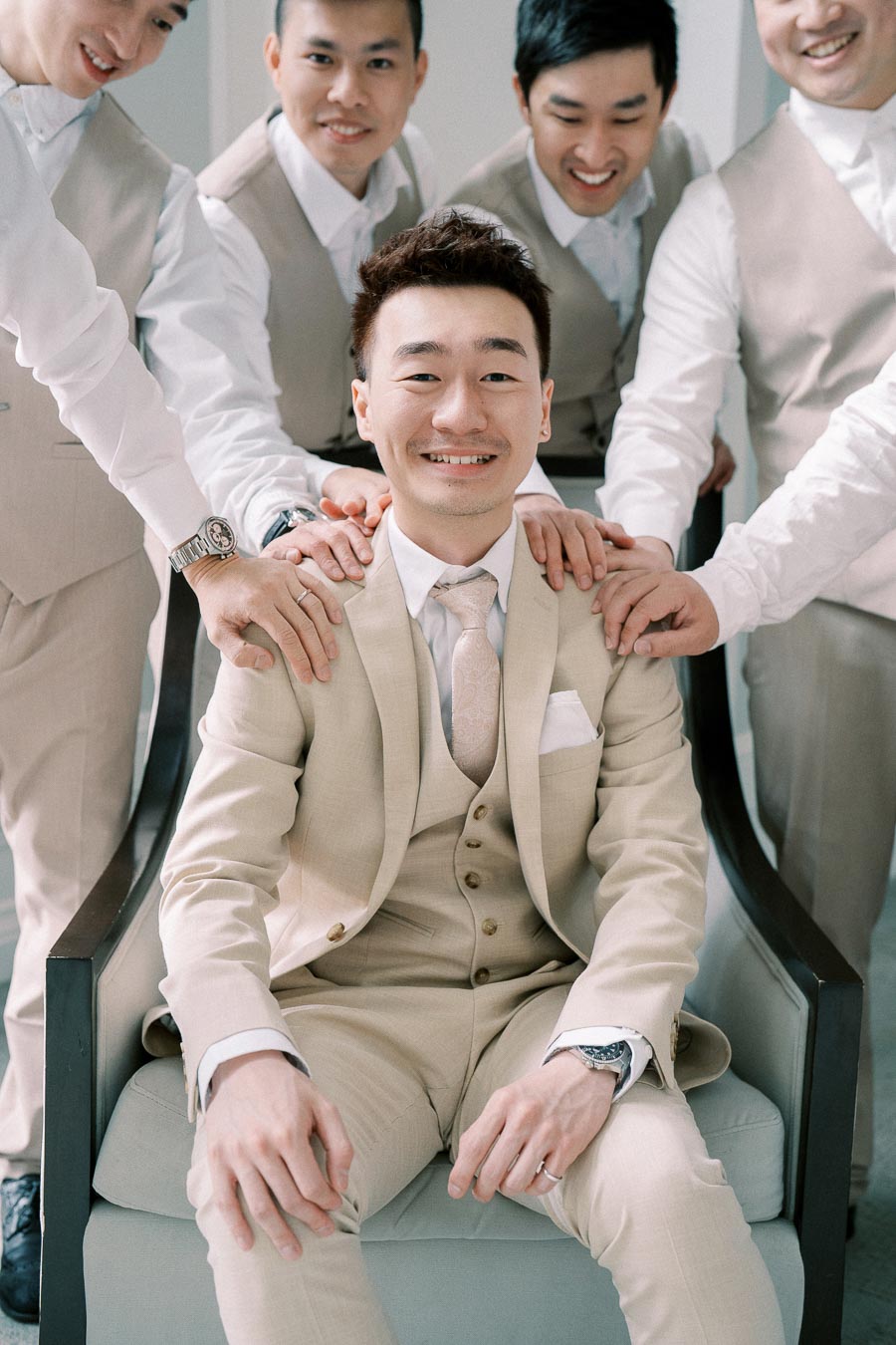 Group of smiling groomsmen in beige suits, gathered around a seated groom on a wedding day, showcasing camaraderie and formal attire.