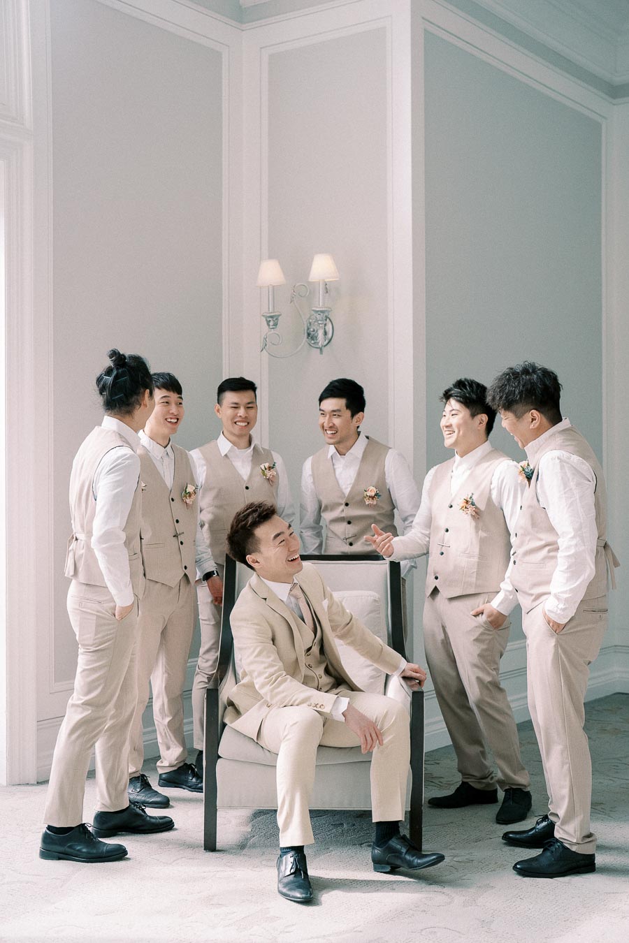 Group of groomsmen in beige suits laughing together in a bright room.