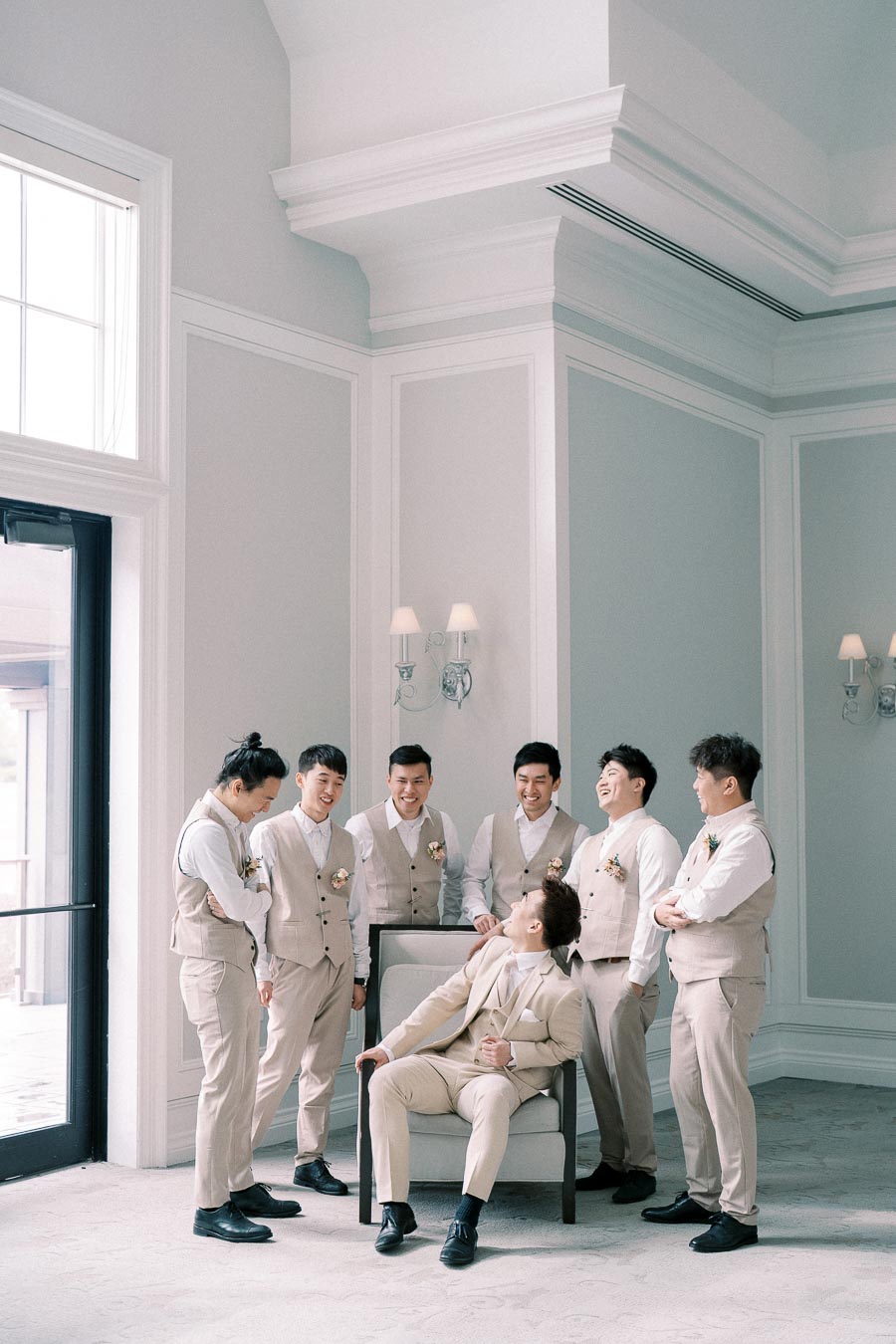 Groom and groomsmen laughing and interacting in a stylish, light-filled room; all dressed in matching beige suits with floral boutonnieres, creating a joyful and elegant pre-wedding moment.