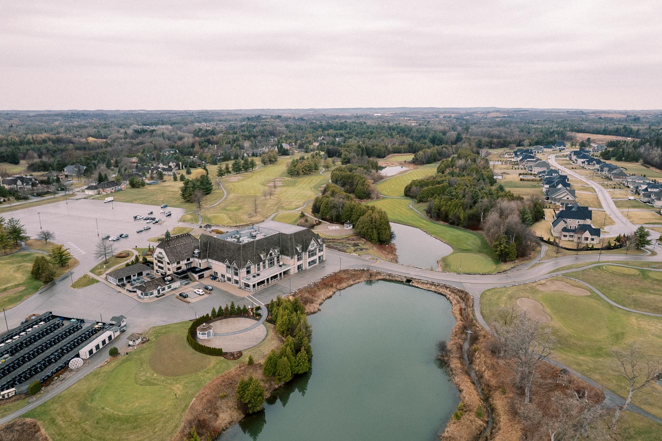 Aerial view of a luxury golf course with clubhouse, surrounded by lush greenery and residential homes, featuring a serene pond and expansive parking area under an overcast sky.