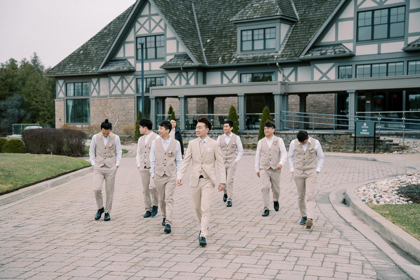 Groomsmen in beige suits and vests walking together outside a Tudor-style building, showcasing elegant wedding attire and a cohesive group dynamic.
