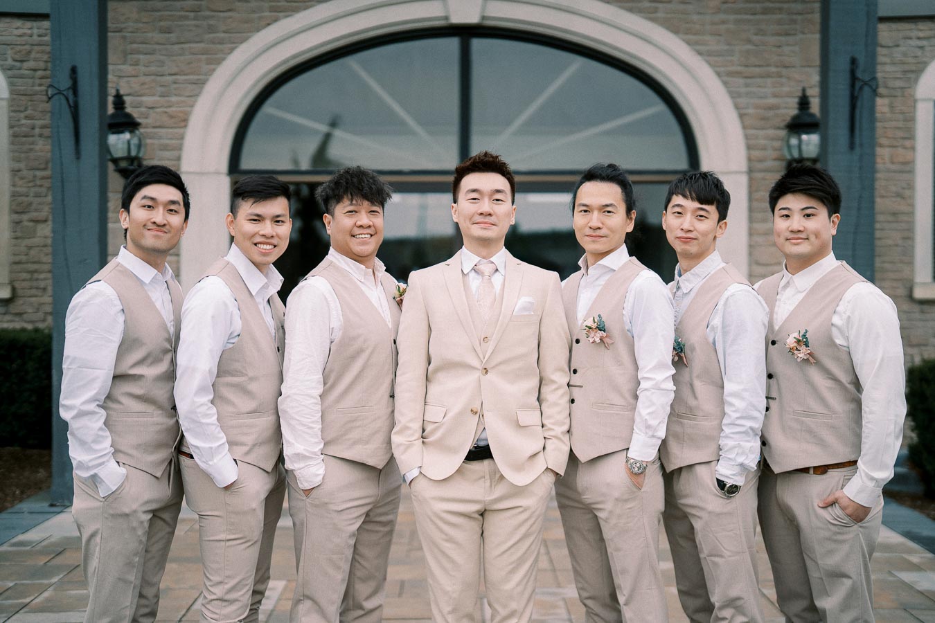 A group of seven groomsmen in beige suits and vests, posing outdoors in front of a classic stone building.