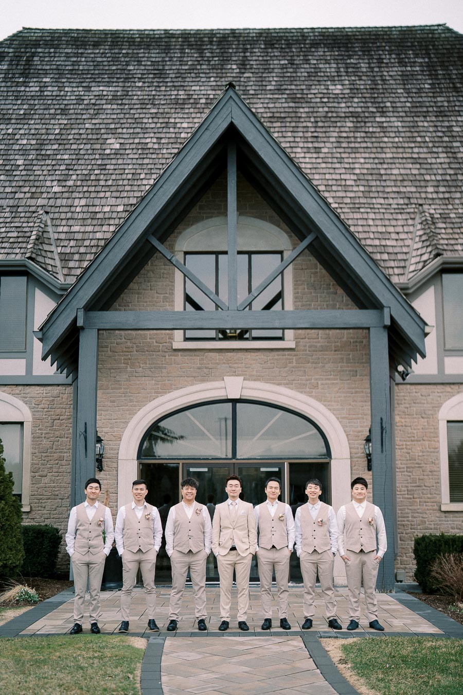 Group of groomsmen in beige suits standing in front of a large stone building with a peaked roof, posing for a wedding photo.