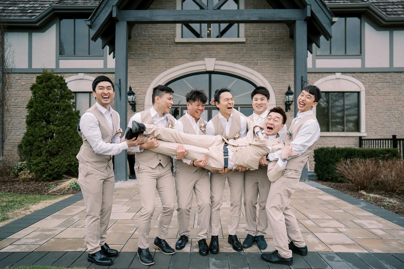 Groomsmen in matching beige suits joyfully lift the groom in front of a rustic building, capturing a fun moment at a wedding celebration.