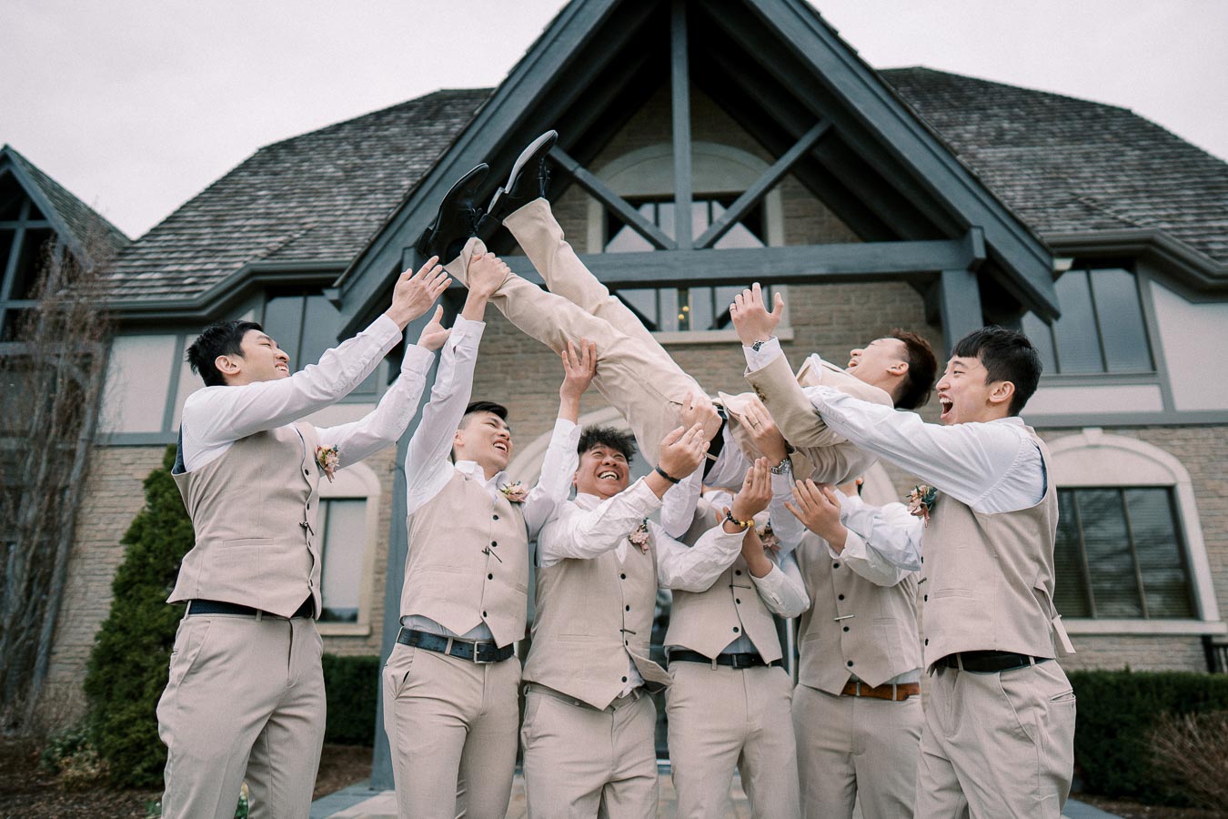Groomsmen in beige vests joyfully tossing a tuxedo-clad man into the air in front of a rustic house, celebrating a wedding day.