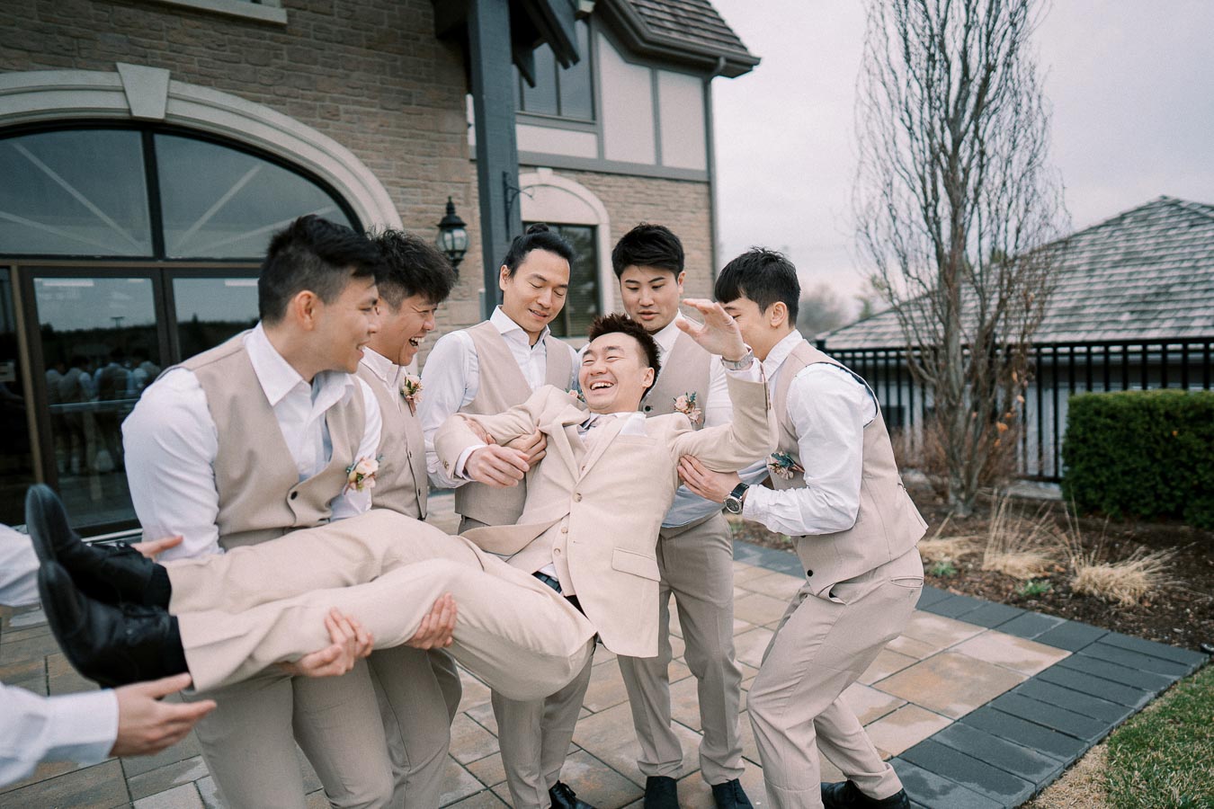 Groom being playfully lifted by groomsmen in tan suits outside a building, capturing a joyful moment on a wedding day.
