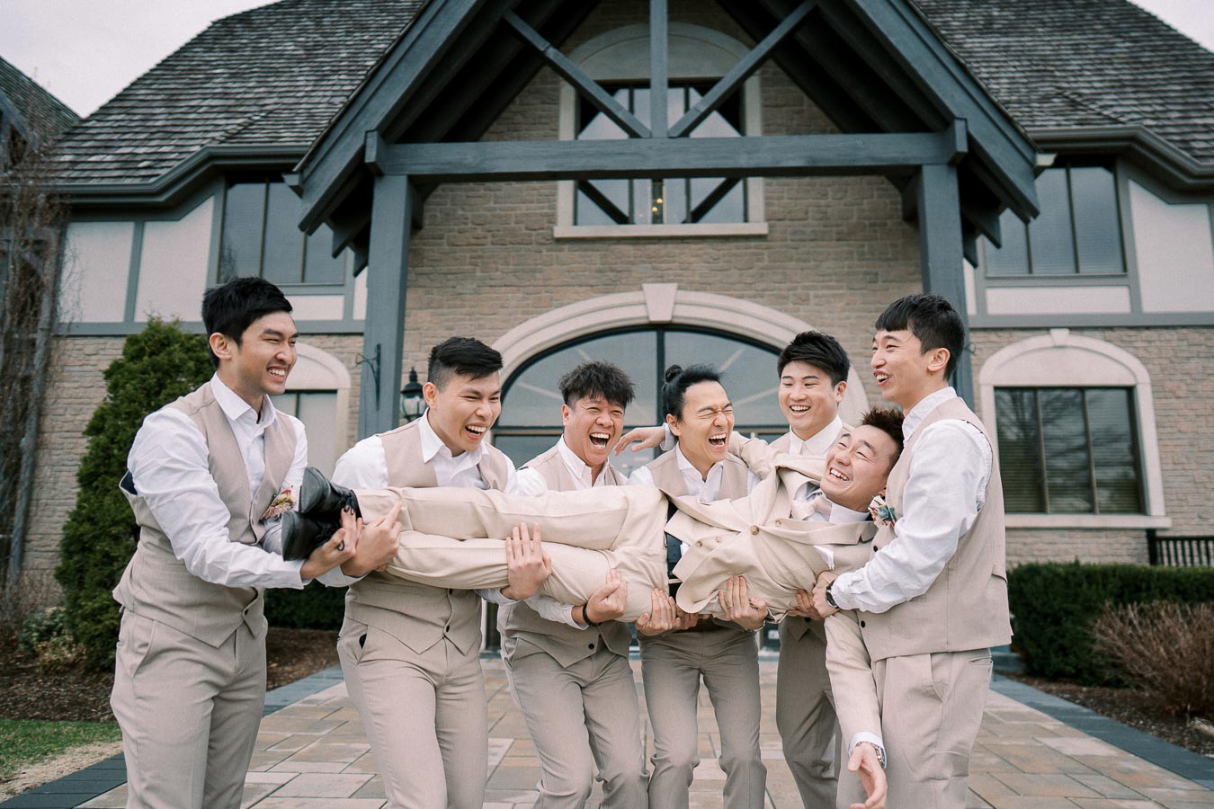 Groomsmen in beige suits joyfully lifting the groom outside a rustic building.